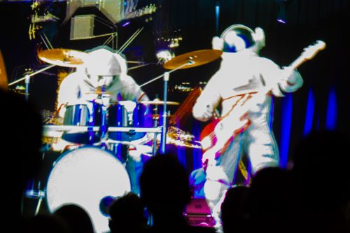 A holographic image of two spacemen in space suits play the drums on the left and an electric guitar on the right